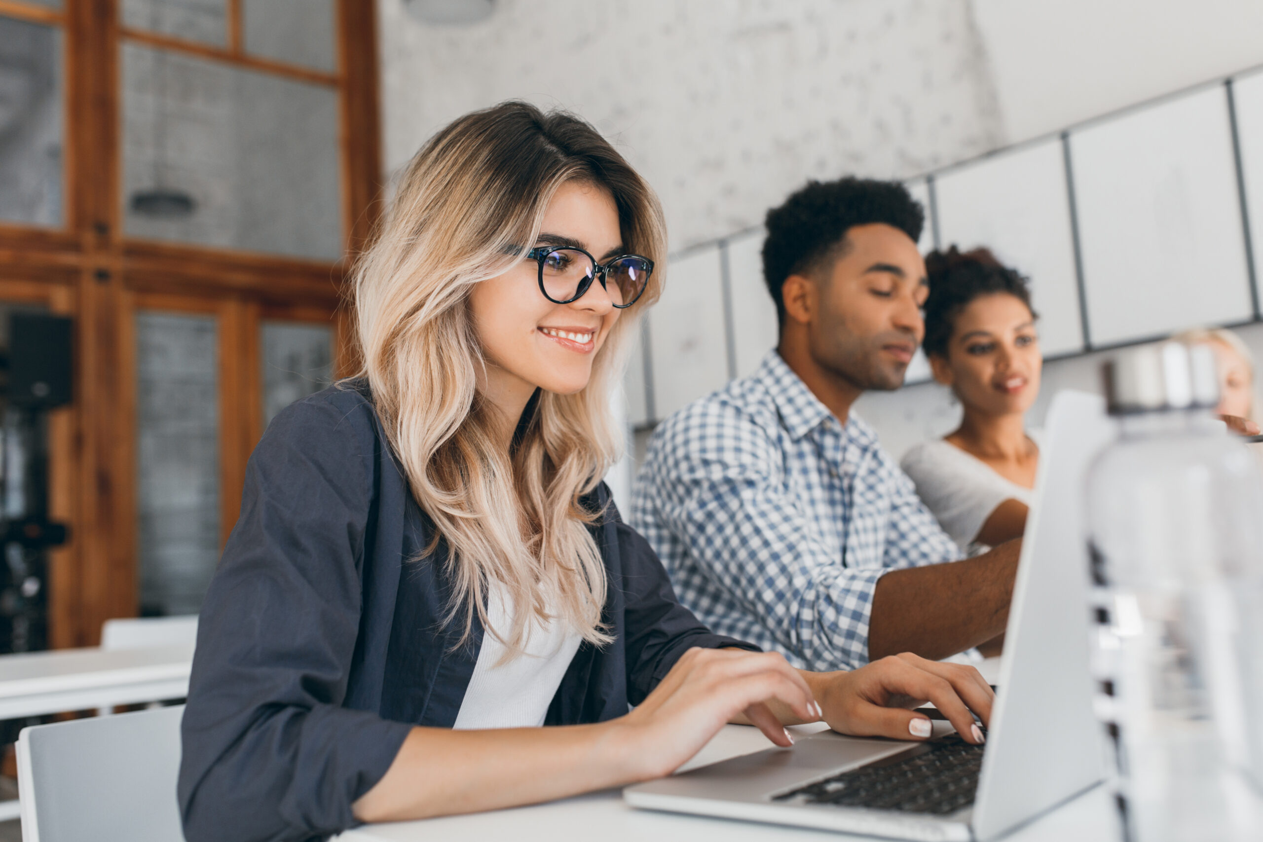 Beautiful curly female freelancer with cute manicure using laptop and smiling. Indoor portrait of blonde secretary sitting beside african coworker in blue shirt..